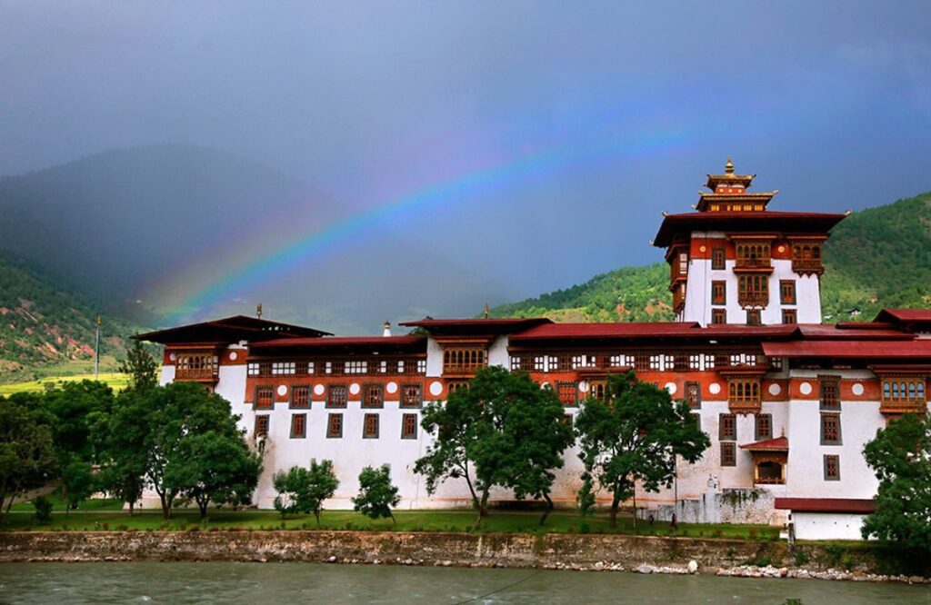 A large whitewashed dzong beneath a rainbow, with forested hills behind