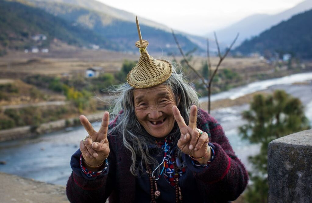 An elderly Bhutanese woman in a bamboo hat smiling beside a river