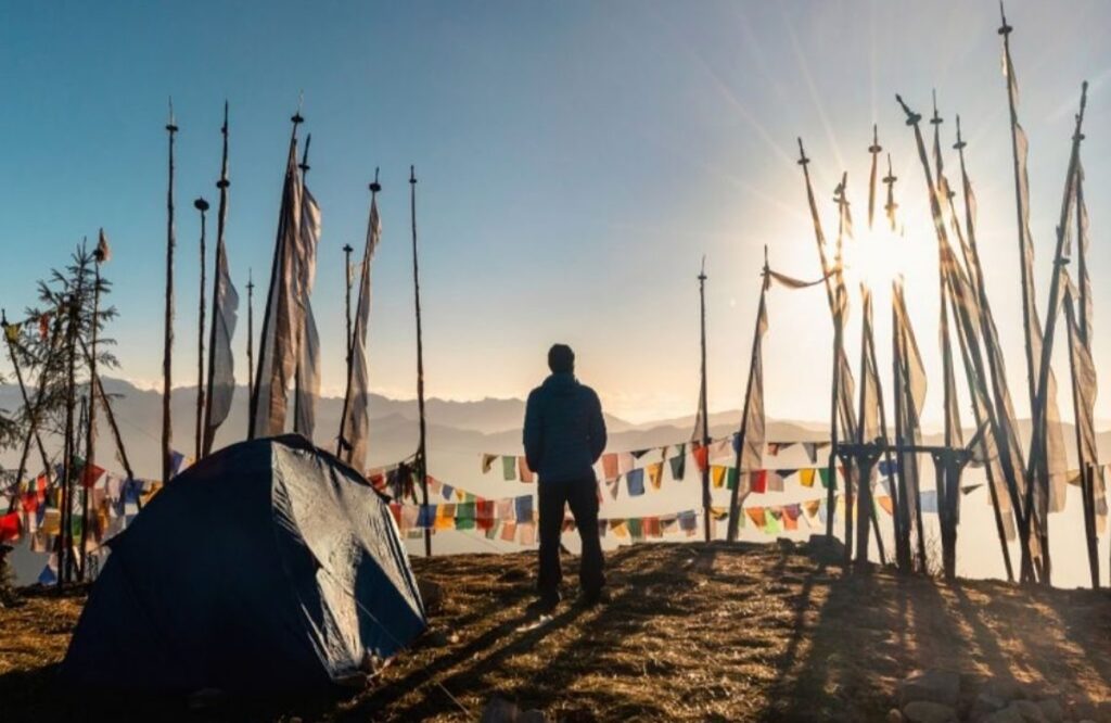 A trekker standing beside a tent at dawn among tall prayer-flag poles