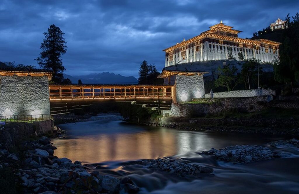 A floodlit dzong and its covered bridge reflected in the river at dusk