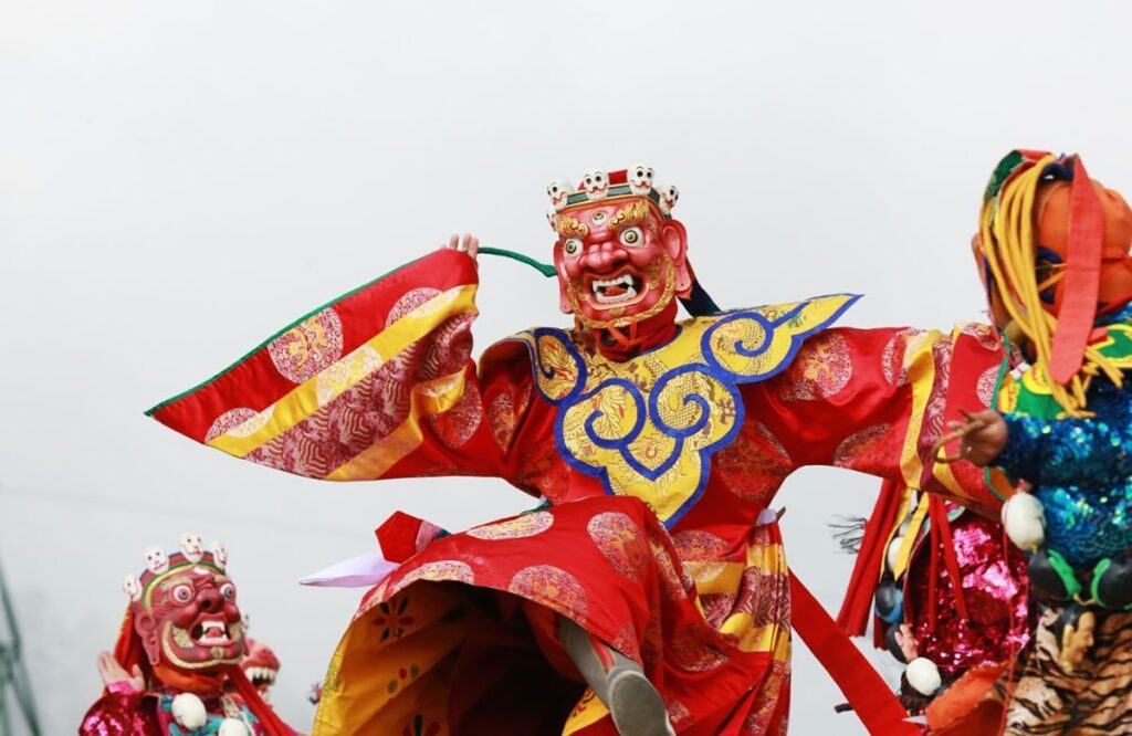 A masked dancer in red and gold silk with arms raised at a Bhutanese festival