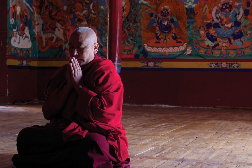 A monk in red robes sitting in meditation on a temple floor before a painted mural