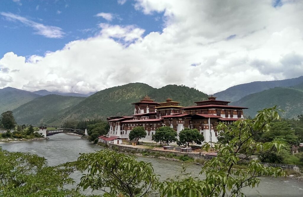 A white dzong beside a wide river at the foot of wooded hills