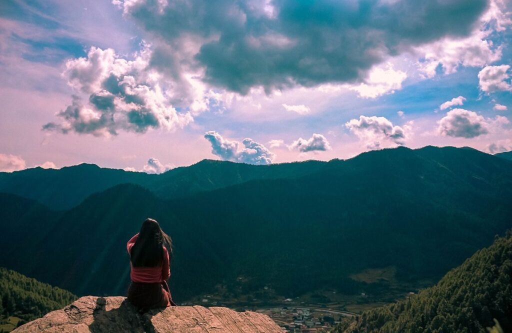 A traveller sitting on a rock watching the sun set over layered forested ridges