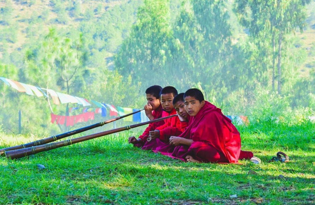 Young monks in red robes sitting on grass beside tall white prayer flags